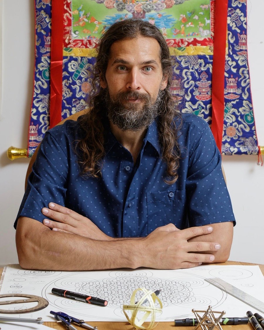 George Leoniak at his drawing table beneath a thangka, a large compass-drawn construction in front of him with dividers, a straightedge and a stellated model